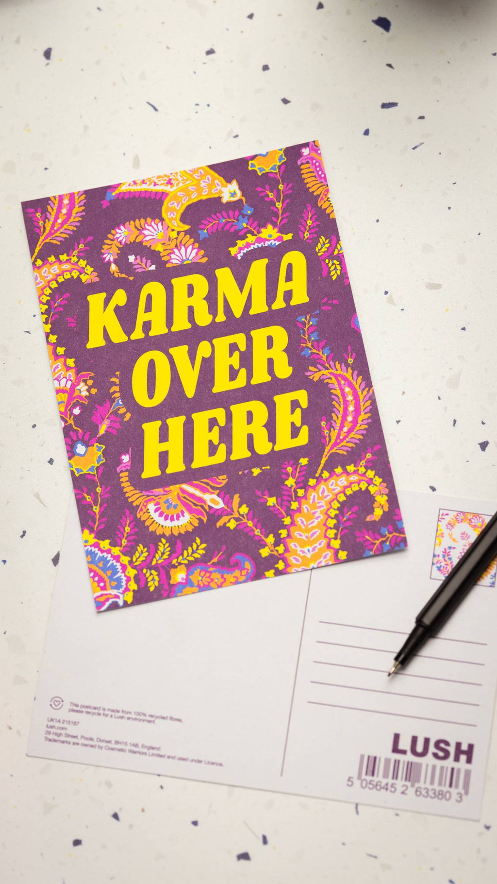 Image shows the colourful Karma Over Here postcard on a table with a blank card and a pen beneath.
