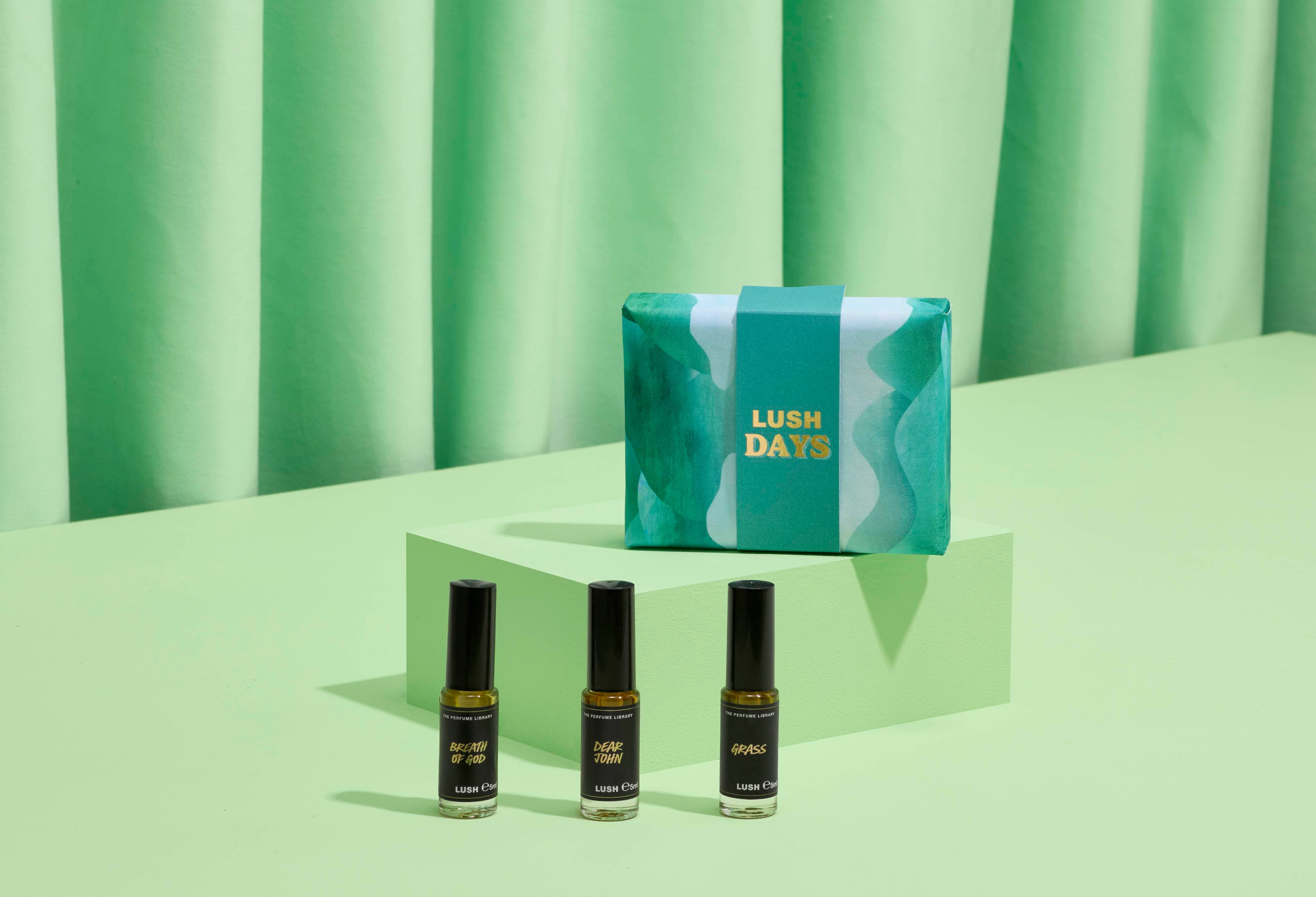 The image shows a "LUSH DAYS" perfume gift set: three thin glass bottles on light green steps. A vibrant teal box with wavy patterns and yellow text sits behind them. A ridged green curtain forms the backdrop.