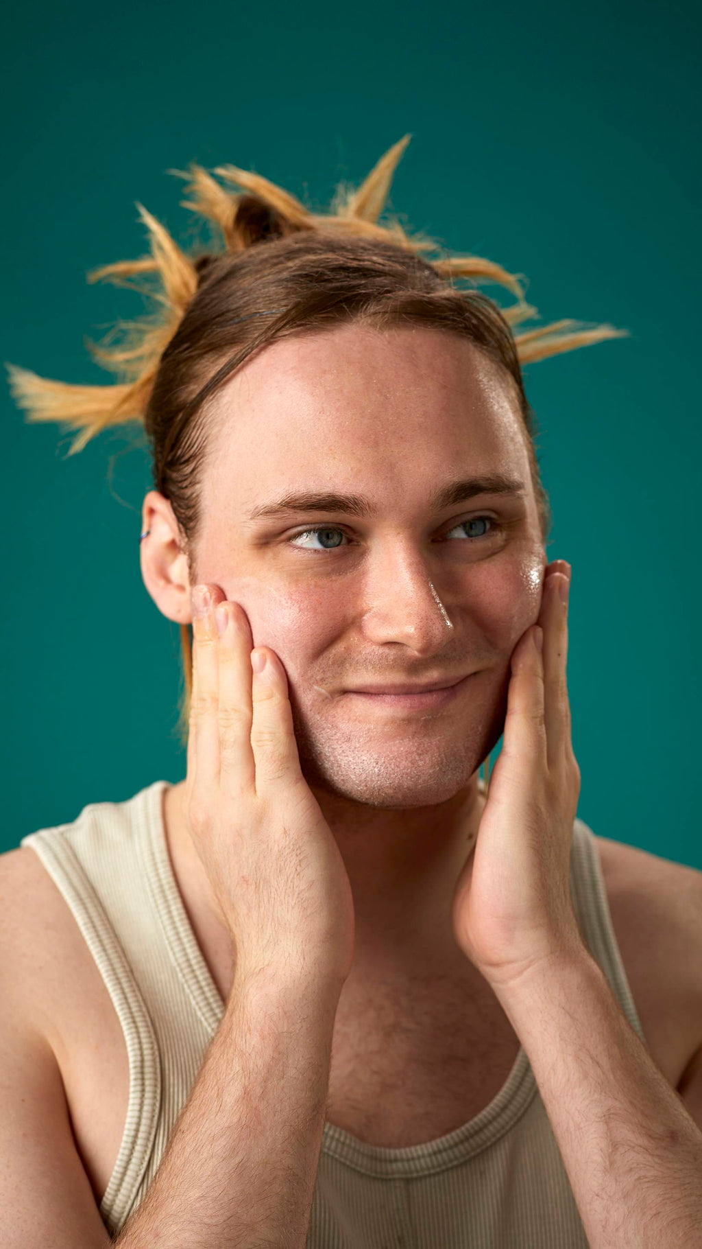 The image shows the model on a teal background with their hair tied back. They have a clean, fresh face as they gently apply the Mostly facial wash over their cheeks.