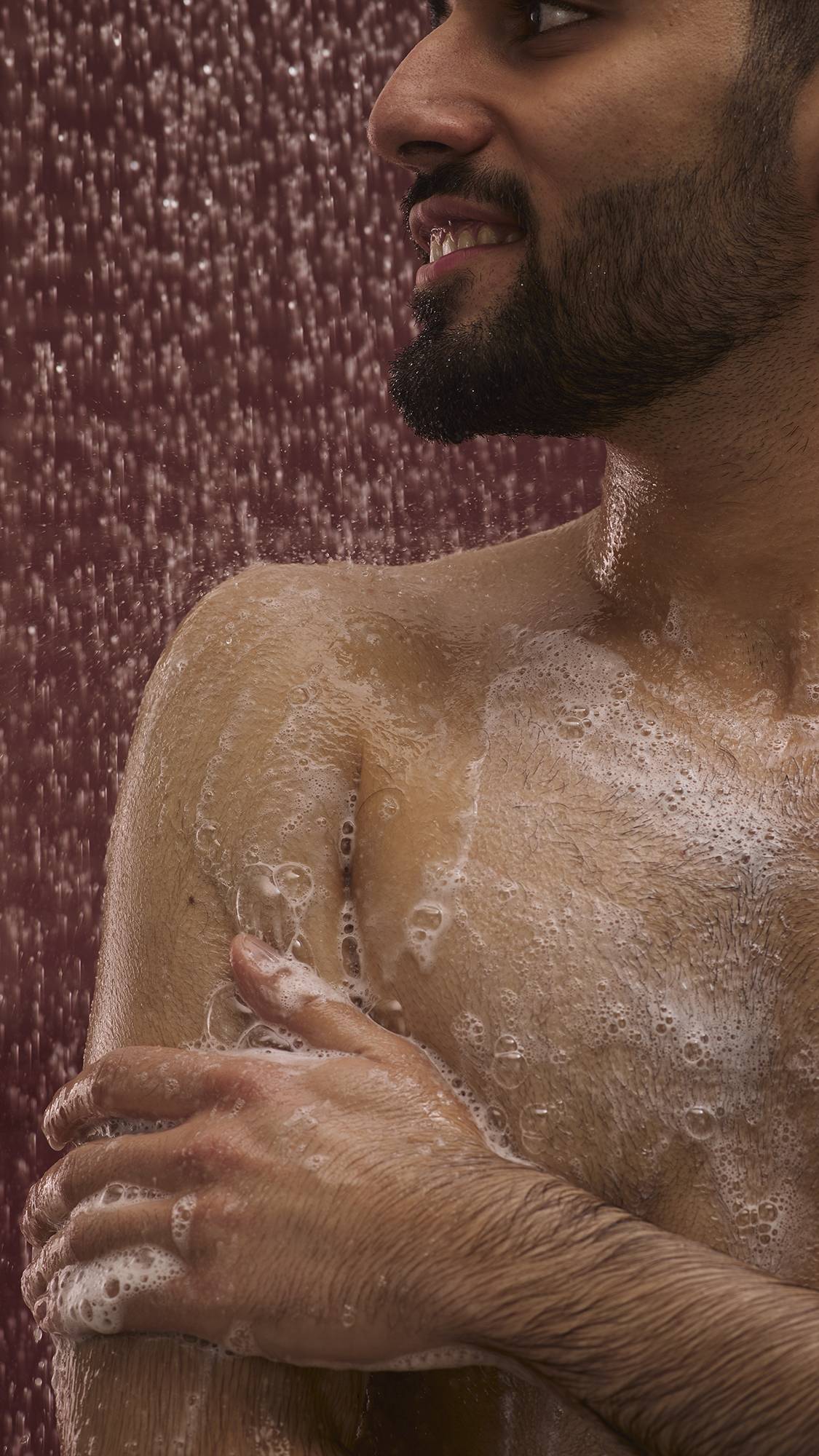The image shows a dark, chocolate-coloured background with falling shower water. In the foreground, the model stands facing the camera as they lather up their shoulder with the glossy Posh Chocolate shower gel to create a soft foam.