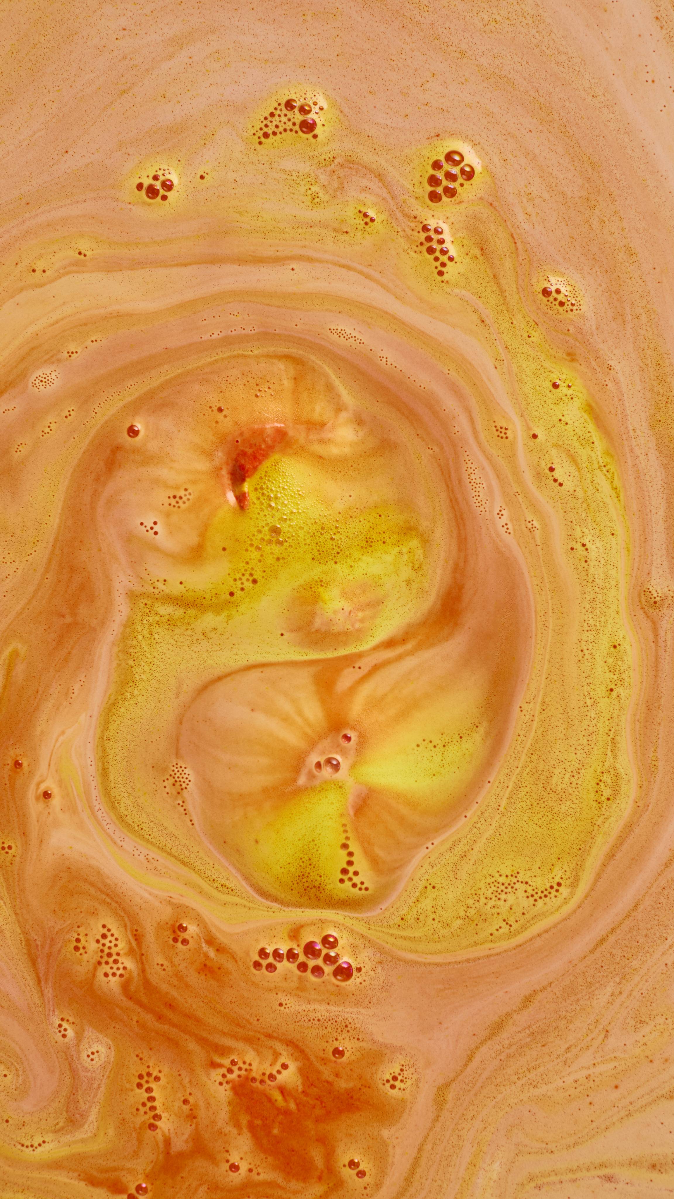 An abstract aerial shot of bath bomb foam swirling in water, creating a large spiral pattern of bright yellow and warm, creamy orange froth. Clusters of fine, orange-tinted soapy bubbles are scattered across the textured surface.