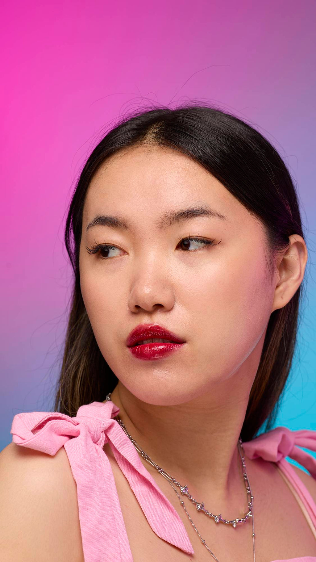 The model has long, straight dark hair and is on a pastel pink, blue and purple hue background. Their make-up has been done with the focal point being the juicy coating of carmine-red tint on their lips created by the Red Maple lip jelly.