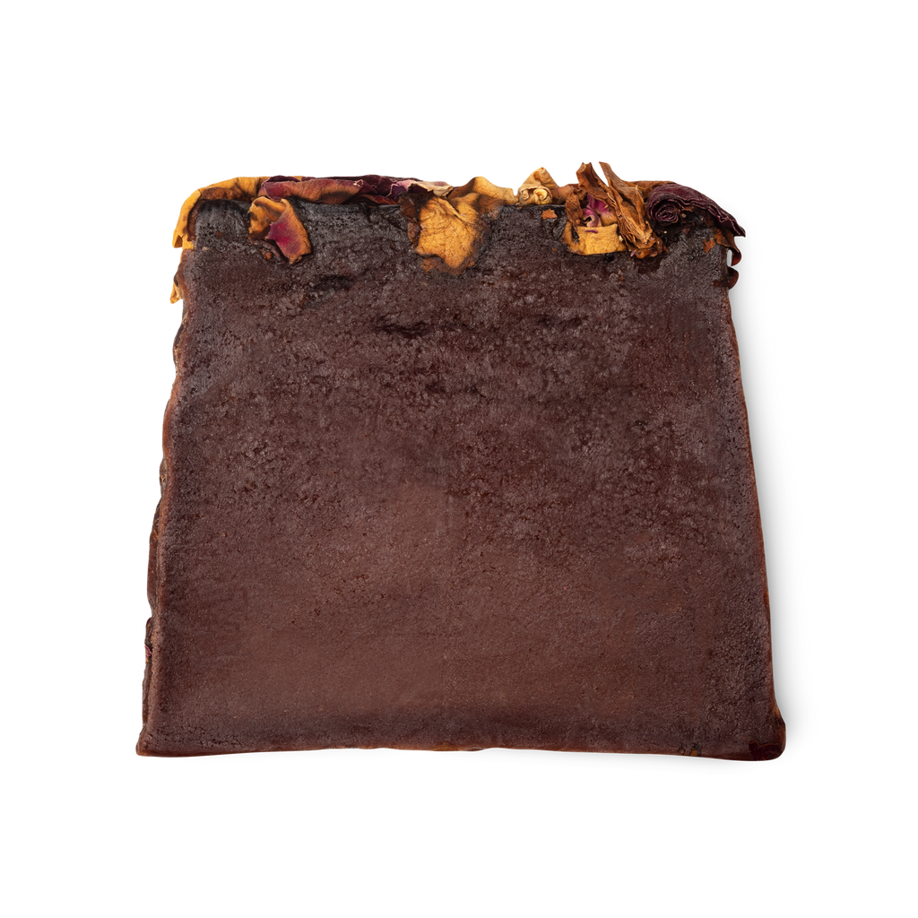 Ro's Argan. A deep purple, moist looking trapezium shaped soap, with a topping of red and yellow rose petals.