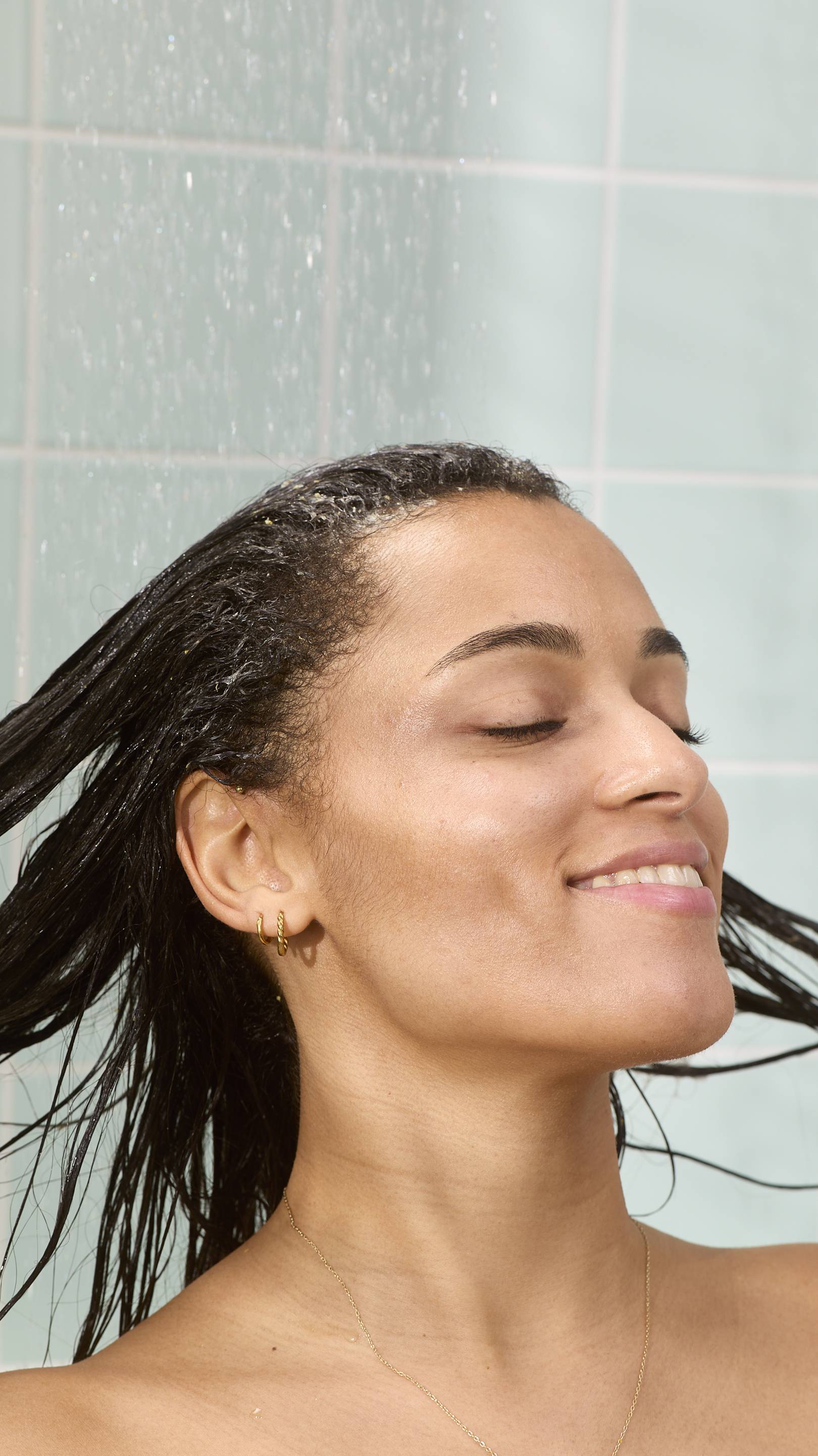 A model is smiling with their eyes closed while rinsing their dark hair under a shower. They are rinsing out the "St Clements" hair treatment product, set against a background of light green bathroom tiles with water droplets visible in the air.