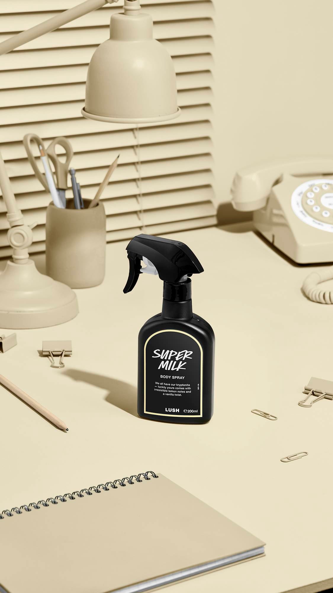 The image shows a black LUSH 'Super Milk' body spray bottle with a pale beige label on a beige desk. Nearby sit a matching desk lamp, a rotary phone, stationery, and a notebook, against window blinds.