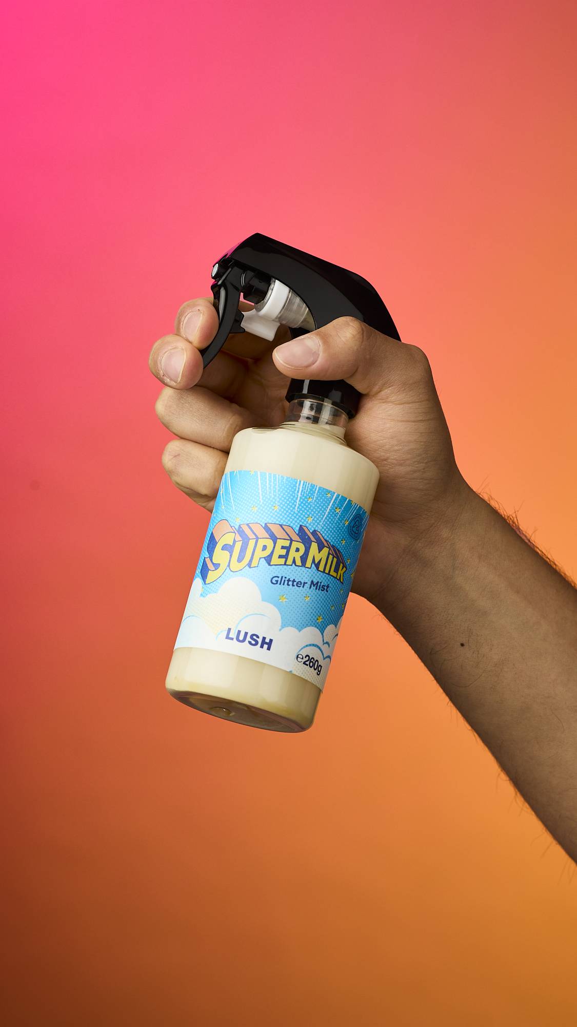A hand holds a bottle of Lush 'Super Milk' Glitter Mist against a vibrant pink and orange gradient background. The bottle contains a cream-coloured liquid and has a bright blue, cloud-patterned label with bold yellow text.