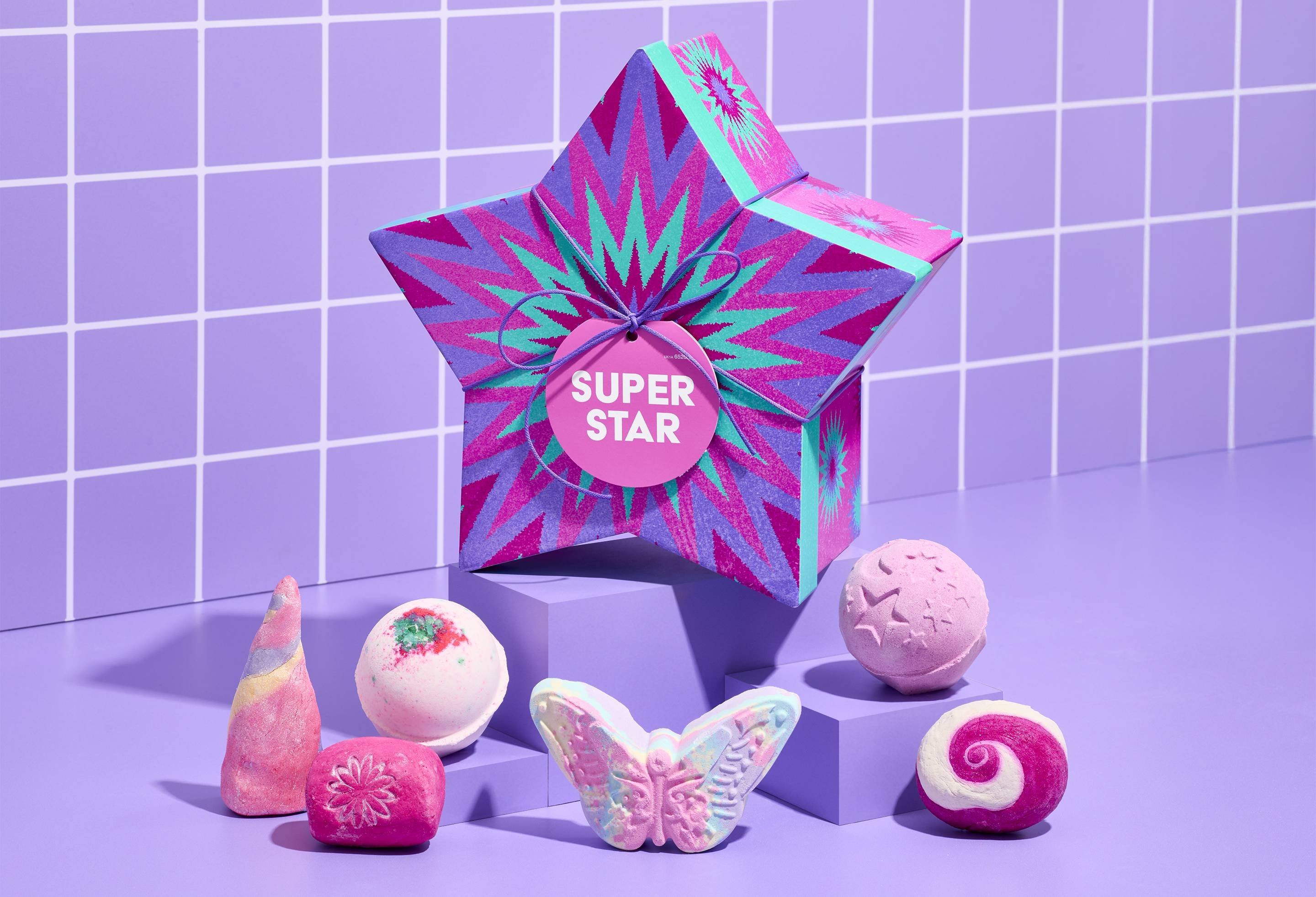 A star-shaped box with purple and turquoise zig-zags. Six colorful bath bombs and bubble bars. Displayed on purple blocks against a matching tiled background.