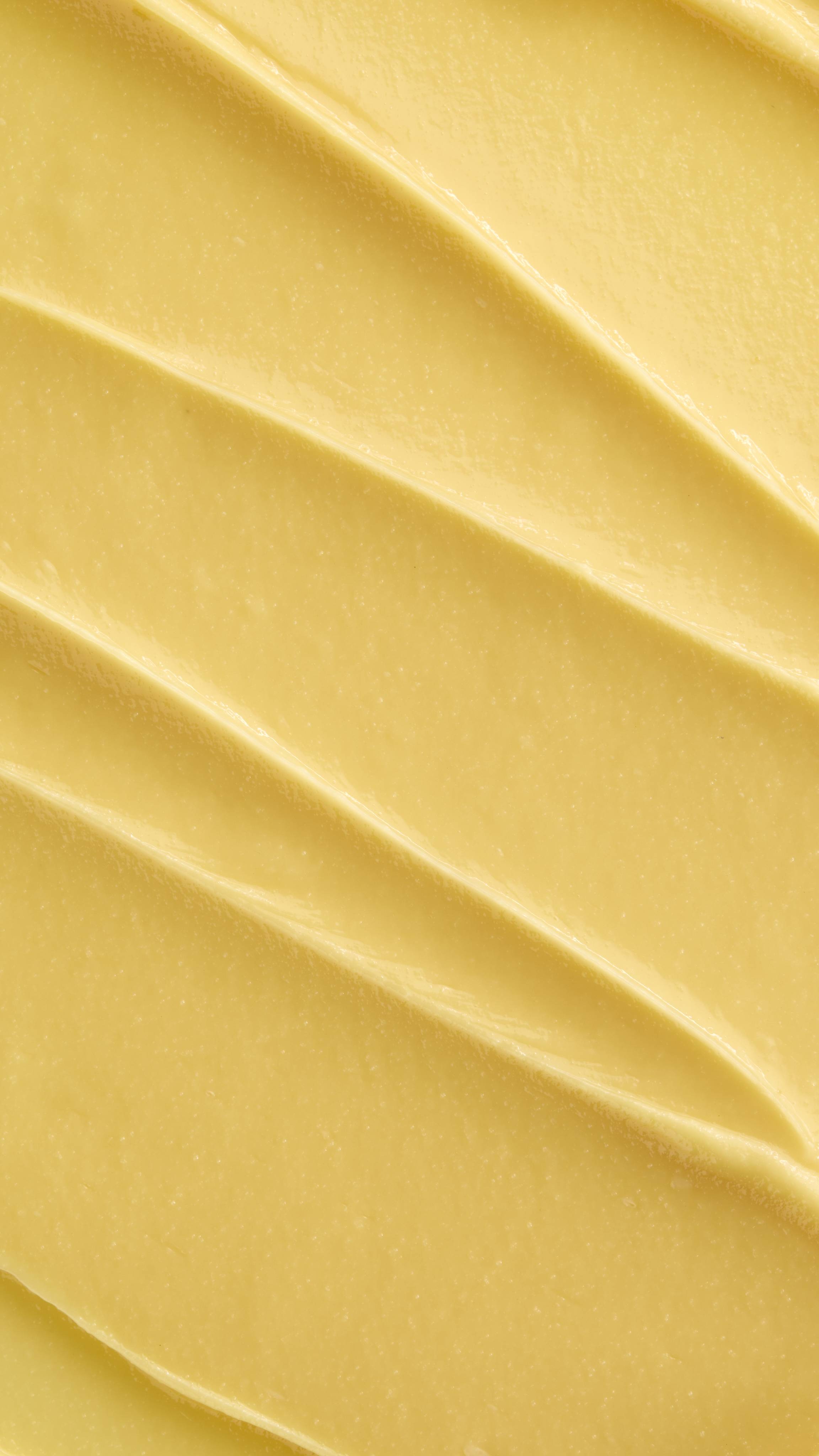 A super close-up image showing diagonal, overlapping smears of the thick, creamy, pale yellow Lush "Turmeric And Argan" facial cleanser. The smooth product has a matte finish and subtle ridged textures.