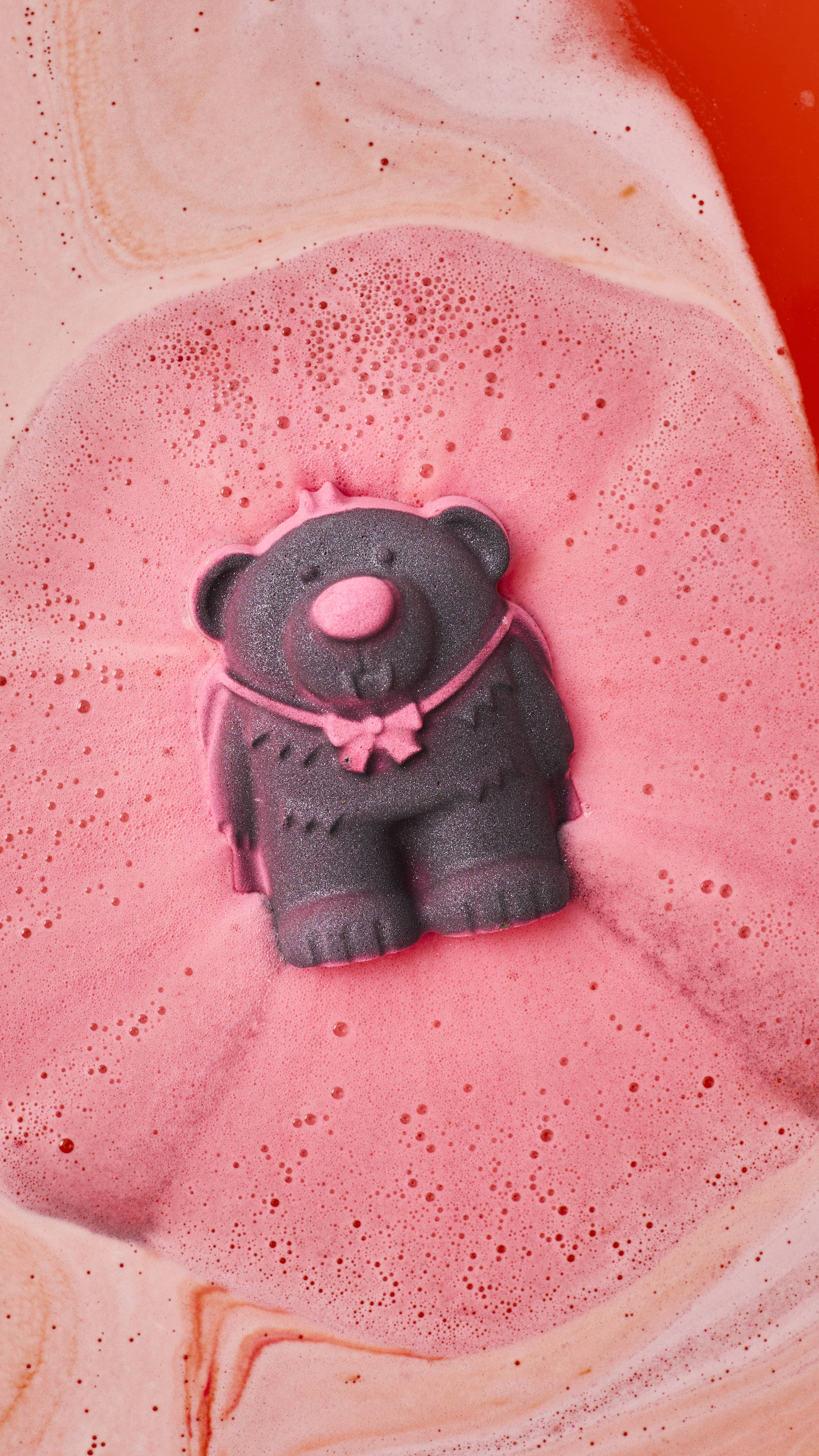 A dark grey vampire bear bath bomb with a pink bow fizzes and dissolves in bathwater, creating a large pool of frothy pink foam surrounded by orange and peach-coloured swirls.